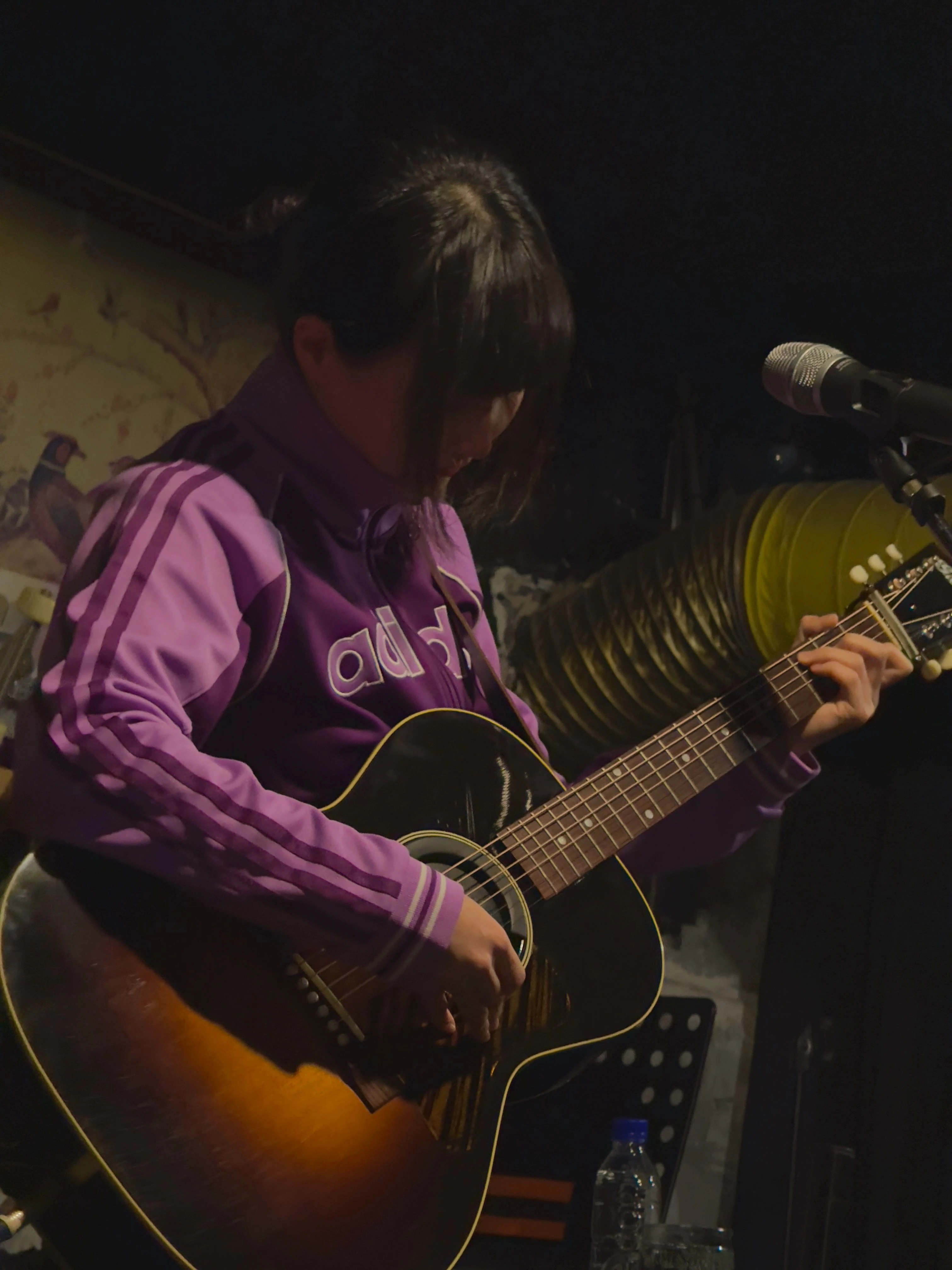 트레이닝 복을 입은 가수 정우가 마이크 스탠드 앞에서 어쿠스틱 기타를 연주하고 있다.