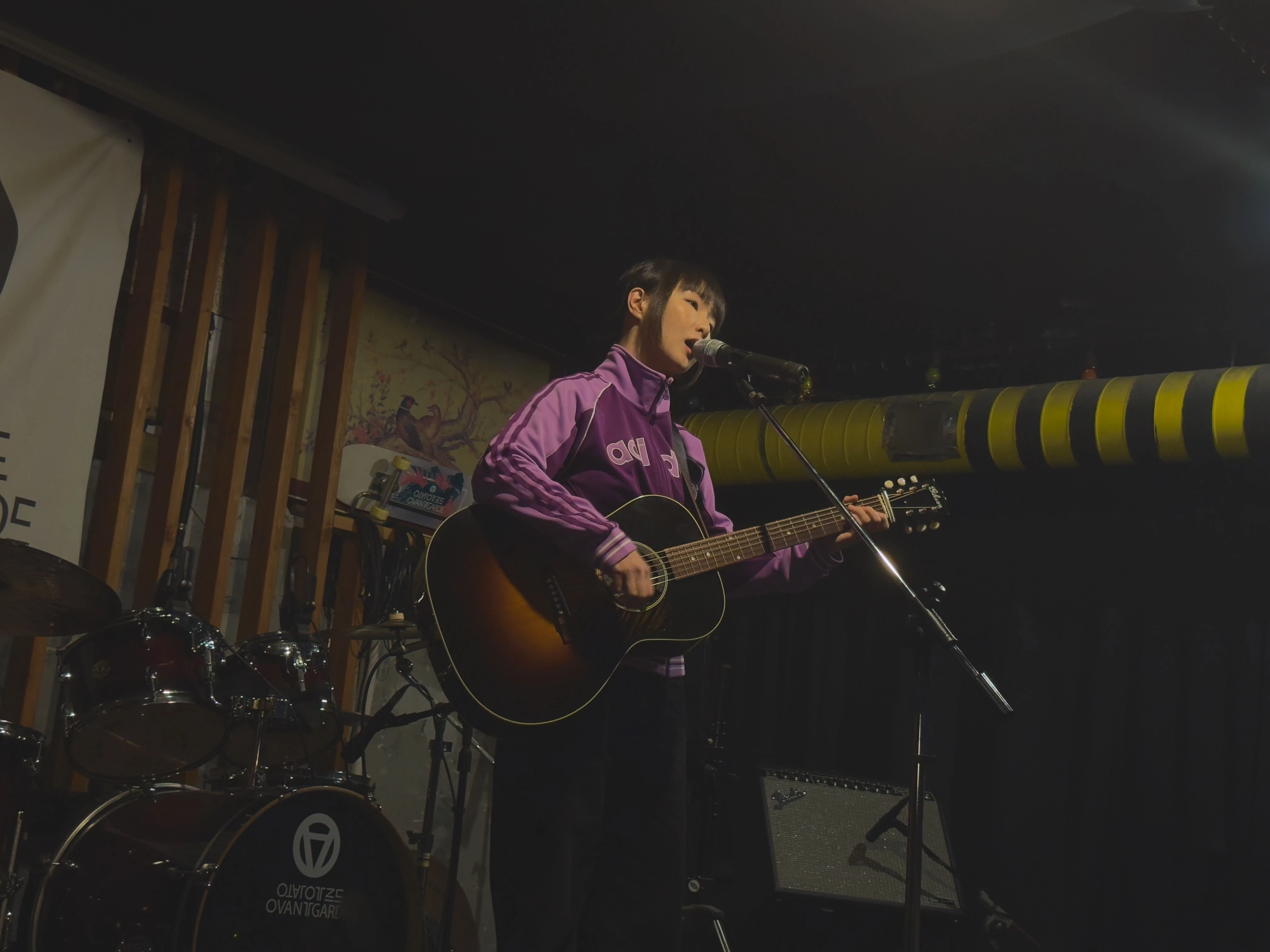 트레이닝 복을 입은 가수 정우가 마이크 스탠드 앞에서 어쿠스틱 기타를 연주하며 노래를 부르고 있다.