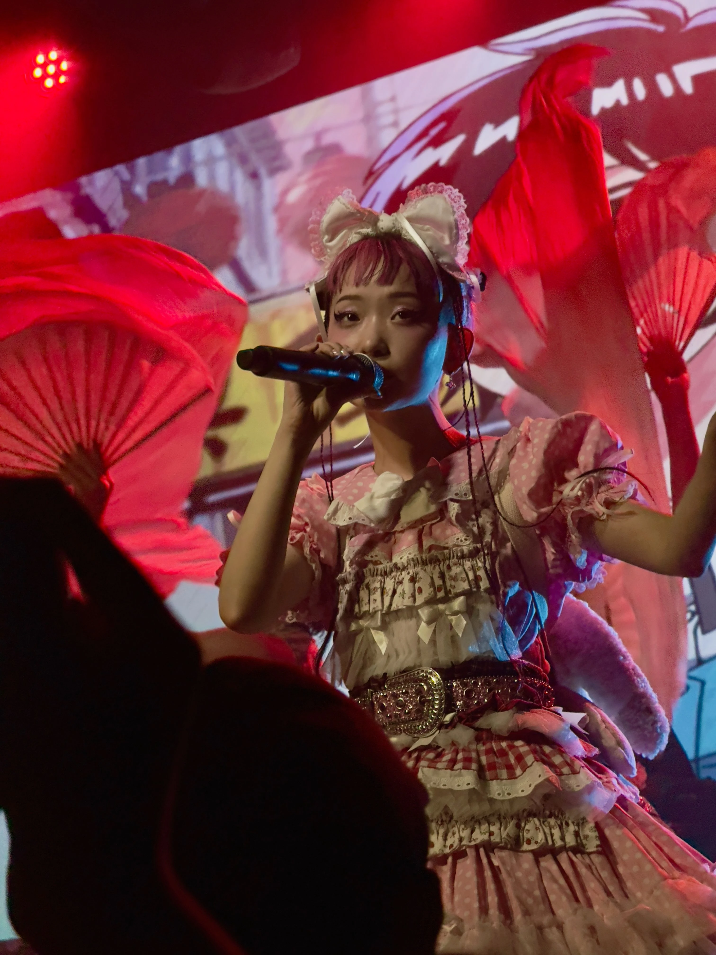 롤리타 패션 복장을 한 WEDNESDAY CAMPANELLA(수요일의 캄파넬라)의 우타하가 마이크를 잡고 노래를 부르고 있다. 무대의 댄서들은 부채춤을 추고 있고, 무대 뒤 디스플레이에는 배경 애니메이션이 표시되고 있다.