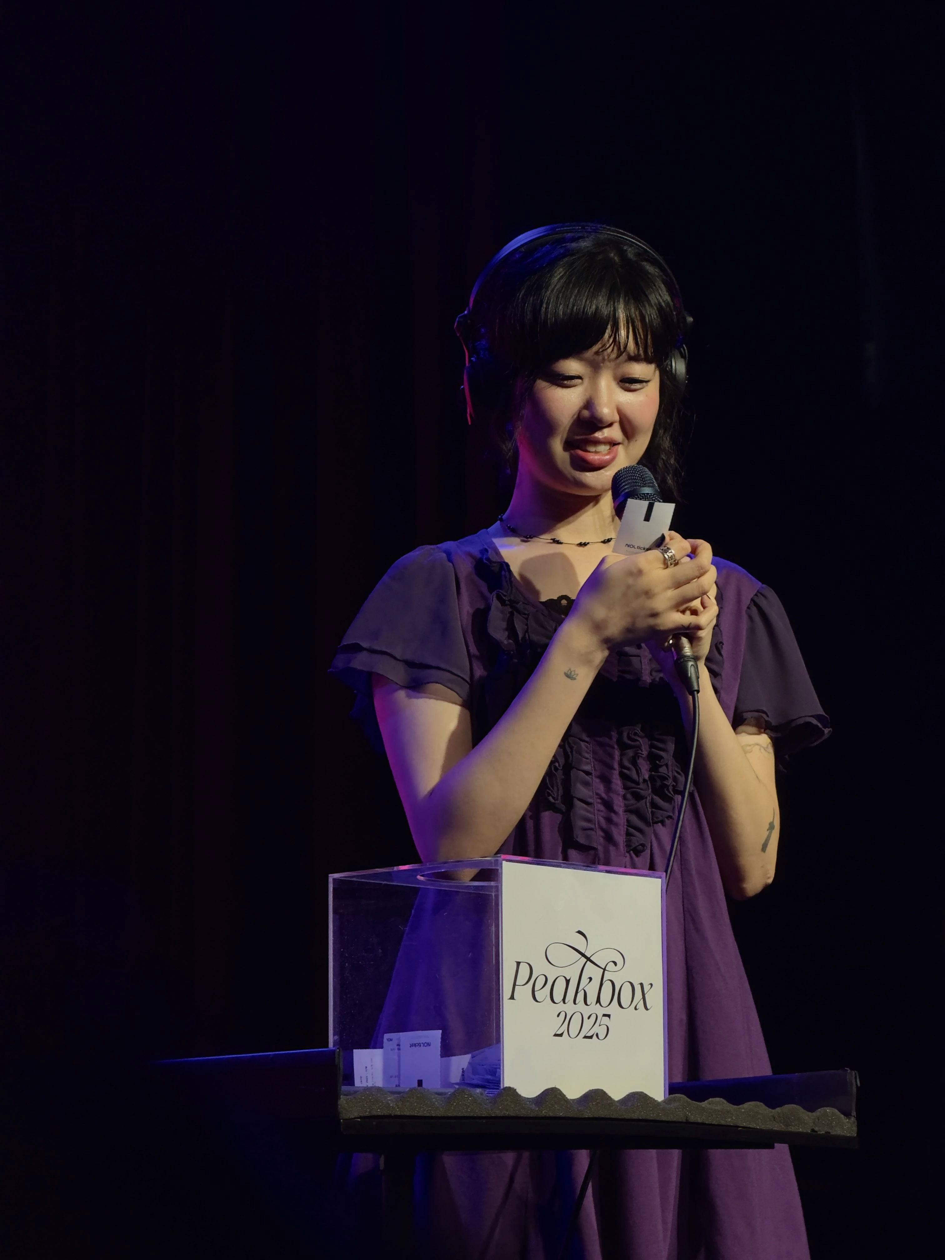 윤지영이 Peakbox 2025라고 적힌 아크릴 상자 안에서 관객들의 회수용 티켓 중 하나를 꺼내고 웃으며 바라보고 있다.