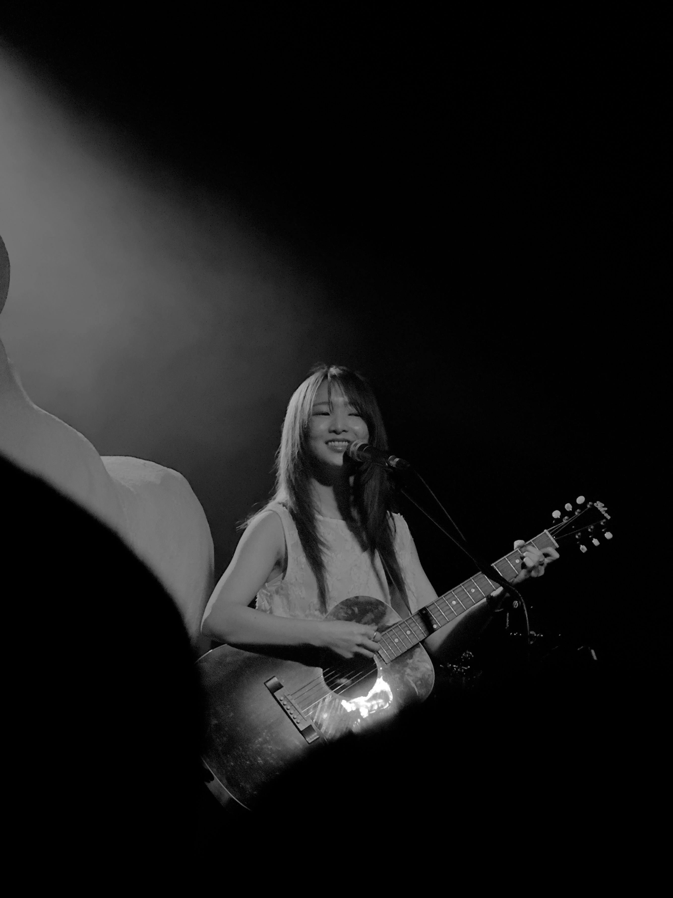 정우가 어쿠스틱 기타를 연주하면서 마이크 스탠드 앞에서 미소를 지으며 노래를 부르고 있다.