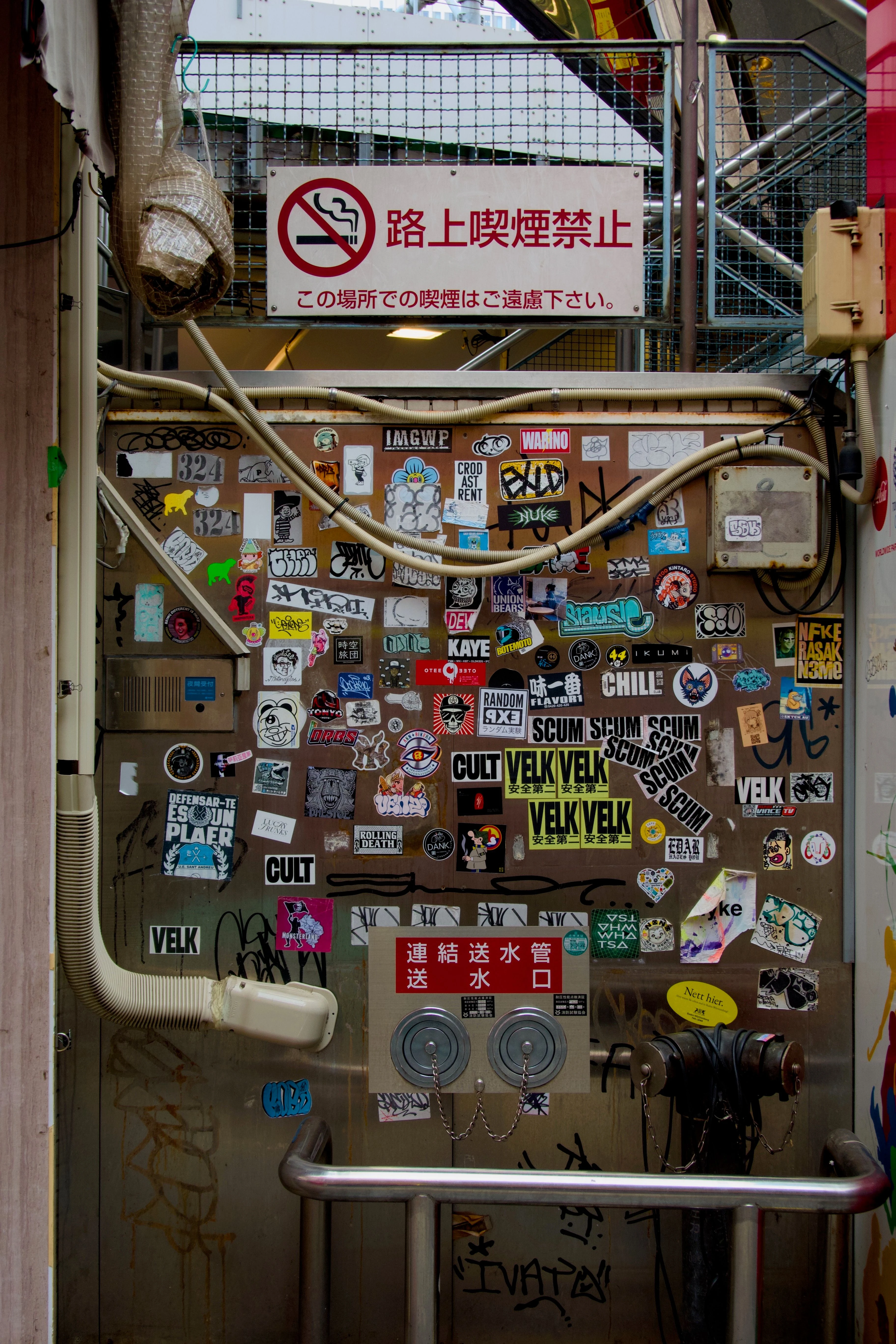 흡연 금지 표지판 아래에 있는 철 재질의 벽에 여러 스티커들이 붙어 있다.