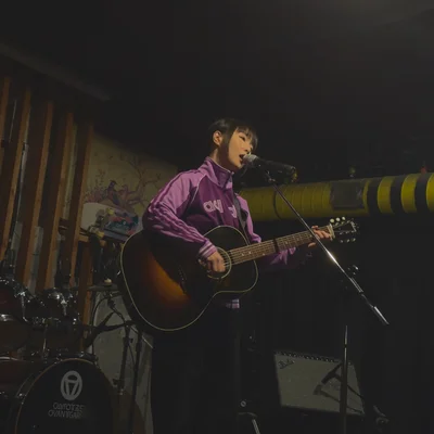 트레이닝 복을 입은 가수 정우가 마이크 스탠드 앞에서 어쿠스틱 기타를 연주하며 노래를 부르고 있다.