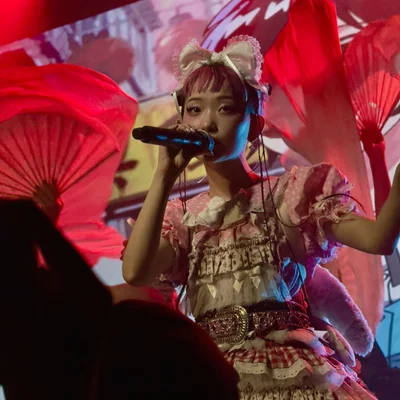 롤리타 패션 복장을 한 WEDNESDAY CAMPANELLA(수요일의 캄파넬라)의 우타하가 마이크를 잡고 노래를 부르고 있다. 무대의 댄서들은 부채춤을 추고 있고, 무대 뒤 디스플레이에는 배경 애니메이션이 표시되고 있다.